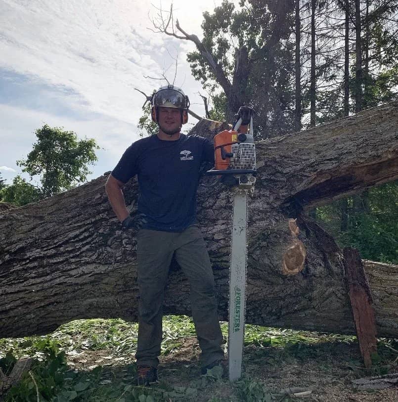 Arborist holding a long-bar chainsaw beside a large fallen trunk.
