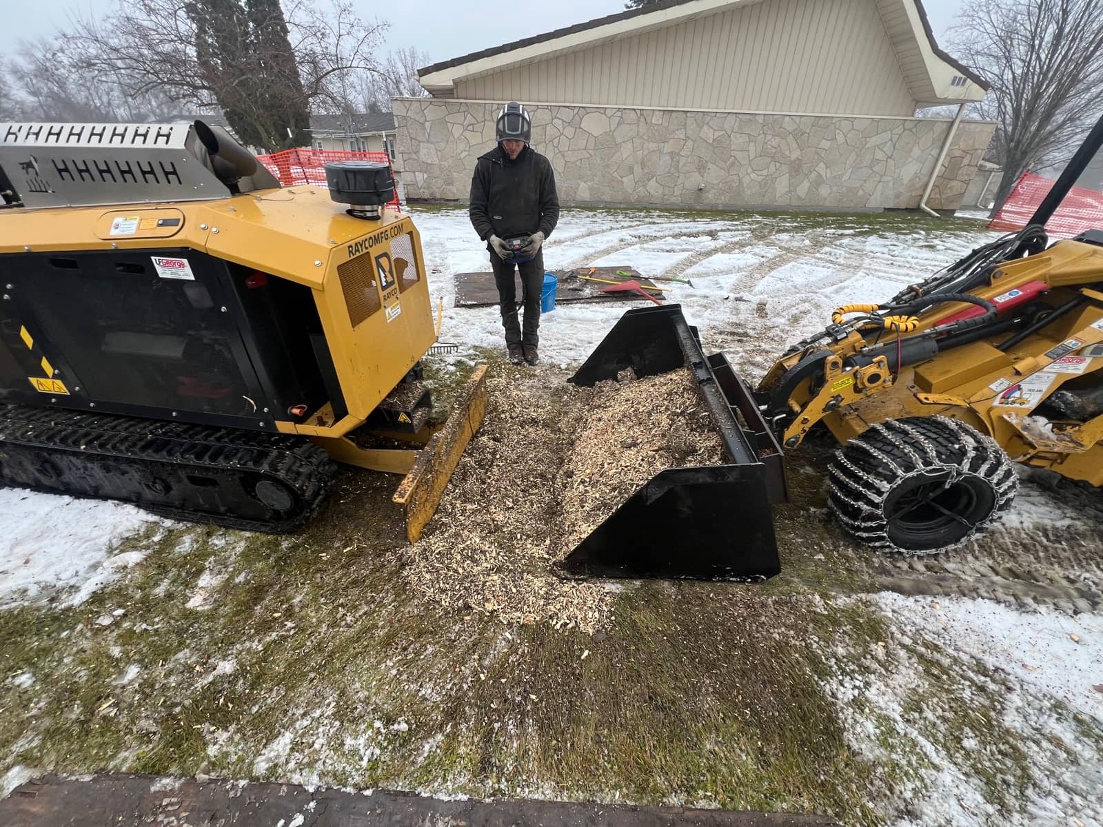 Stump grinder and compact loader beside a pile of wood chips in winter.