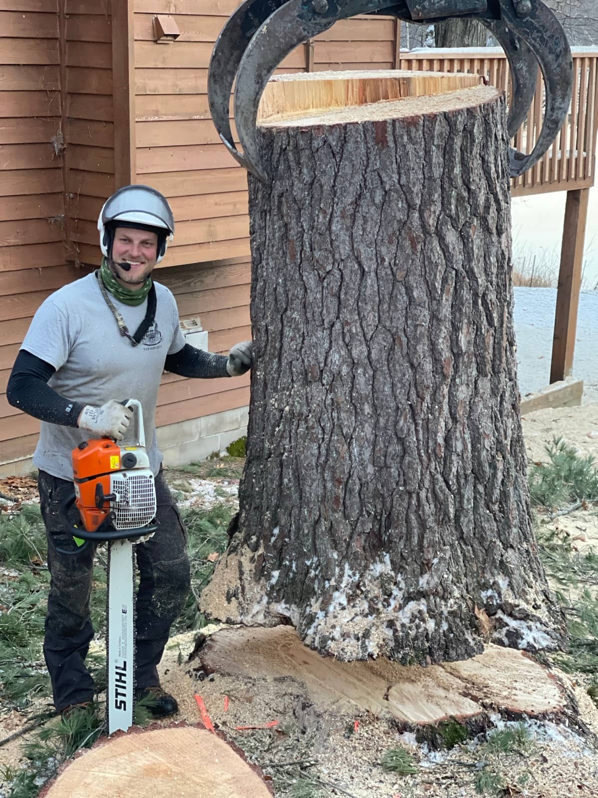 Arborist with a chainsaw beside a trunk held by a grapple.
