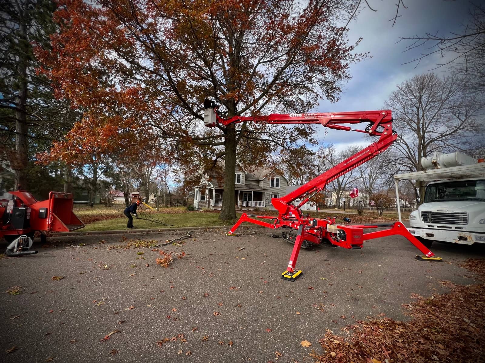 Tracked lift extended into an autumn tree beside work trucks.