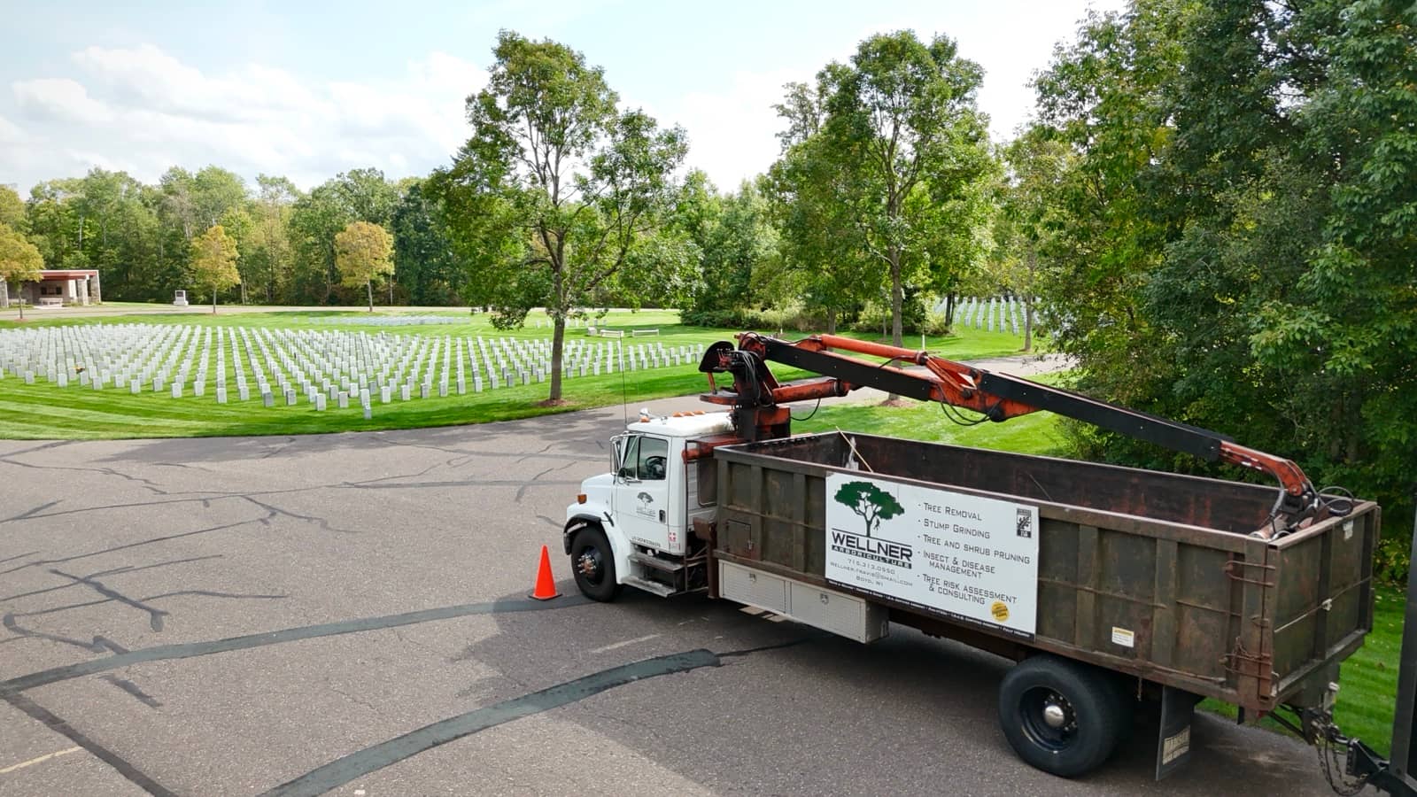 Wellner log truck with a grapple crane beside cemetery grounds.