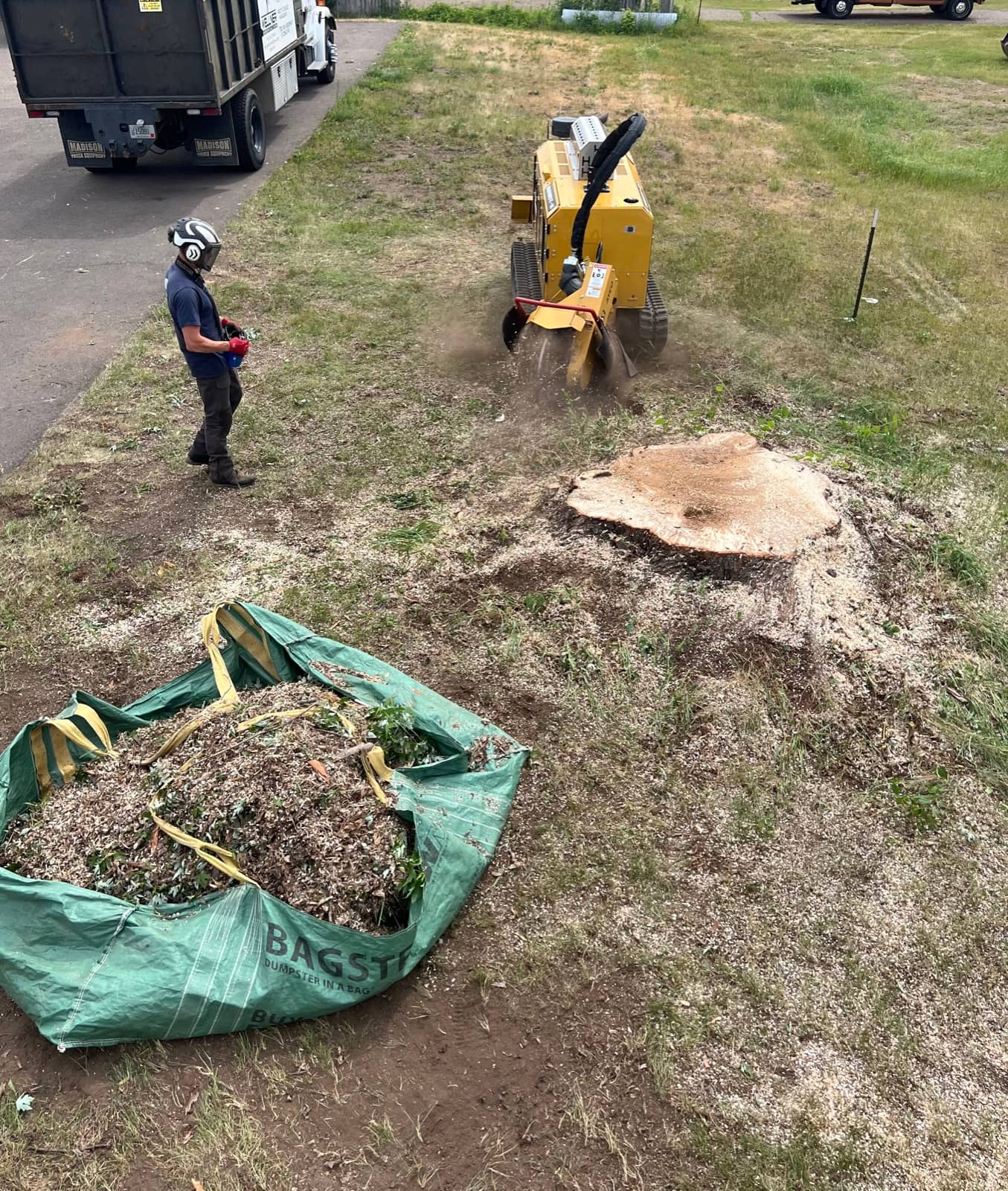 Worker operating a stump grinder beside a stump and a tarp of debris.